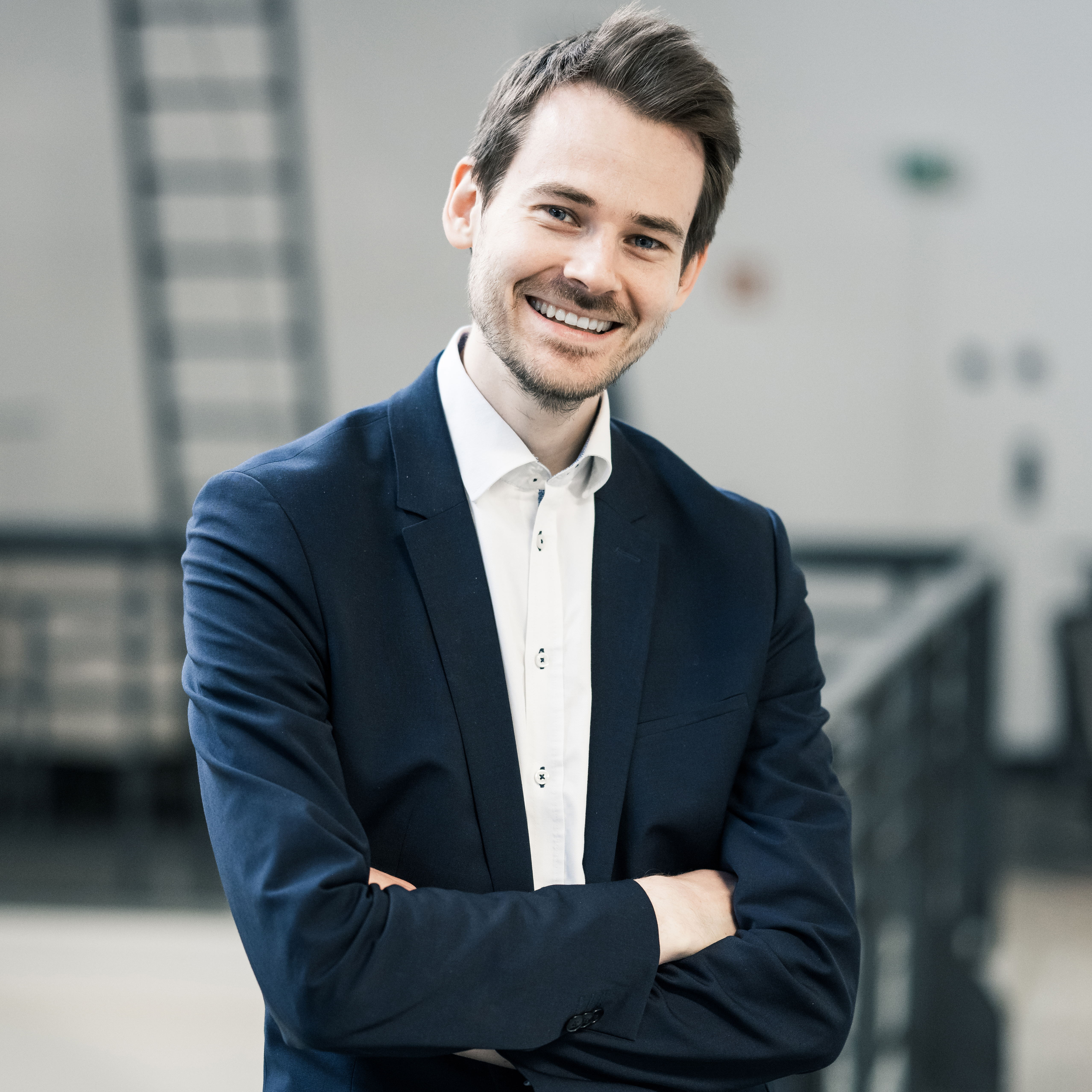 RA René Scheier - Junger Mann lächelt in dunklem Anzug, weißes Hemd, Arme verschränkt, im modernen Innenbereich.