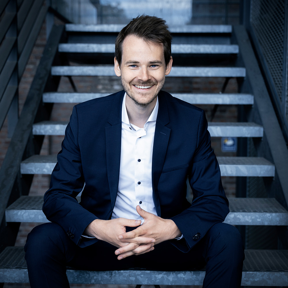 Ein Mann in dunklem Anzug sitzt auf einer Treppe und lächelt freundlich.